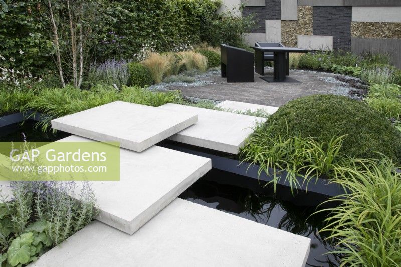 'Shades of Grey' at BBC Gardener's World Live 2021 - paved path over pond leading to seating area in urban contemporary garden, bordered by patchwork hard landscaped wall