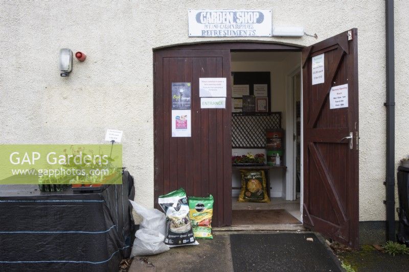 An entrance to a small nursery with some plants on a stall to the left of the entrance door, bags of compost and other sundries beside the door and primroses, log fuel and fish, blood and bone inside the entrance. Spring. 