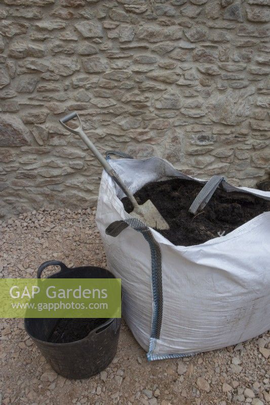 A bulk quantity of compost in a dumpy bag sits beside a plastic trug, half filled with compost with a spade in the dumpy bag. Both sit in front of a newly constructed wall. 
