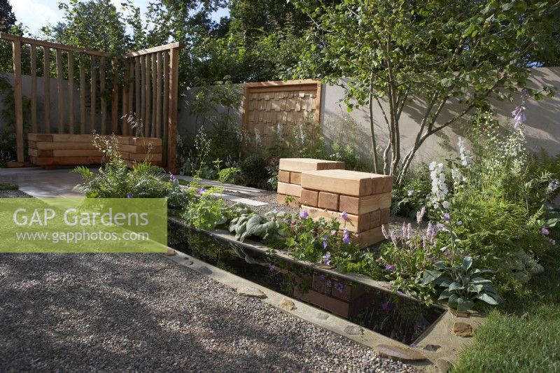 The Communication Garden. Designer: Amelia Bouquet. Water rill with  wooden cubes for seating in the gravelled area and a Corylus avellana tree. RHS Hampton Court Palace Festival 2021.