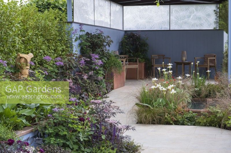 A serene sanctuary with a covered seating area beside a pond planted with Iris 'White Swirl', and surrounded in herbaceous planting in shades of green, purple, bronze and blue.