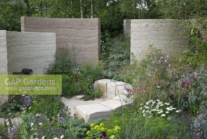 View of a seating area in The Mind Garden with large purbeck stone blocks and opposite, a water feature with ceramic water chutes encased in a clay rendered wall, the meadow planting includes Leucanthemum vulgare, Gladiolus communis subsp. byzantinus, Euphorbia palustris and Stipa gigantea - Designer: Andy Sturgeon - Sponsor: Project Giving Back.