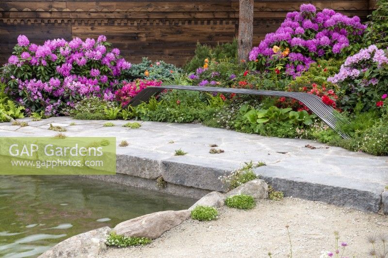 Bespoke iron bench overlooking a pond with colourful Rhododendrons behind - A Swiss Sanctuary, RHS Chelsea Flower Show 2022