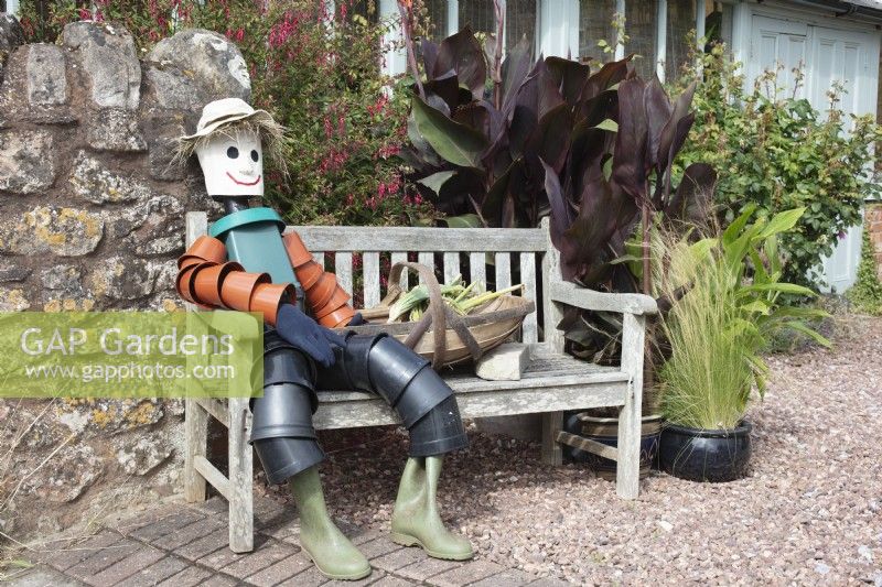 An English village flowershow competition display featuring a scarecrow flowerpot man sitting on a bench next to a basket of homegrown vegetables.