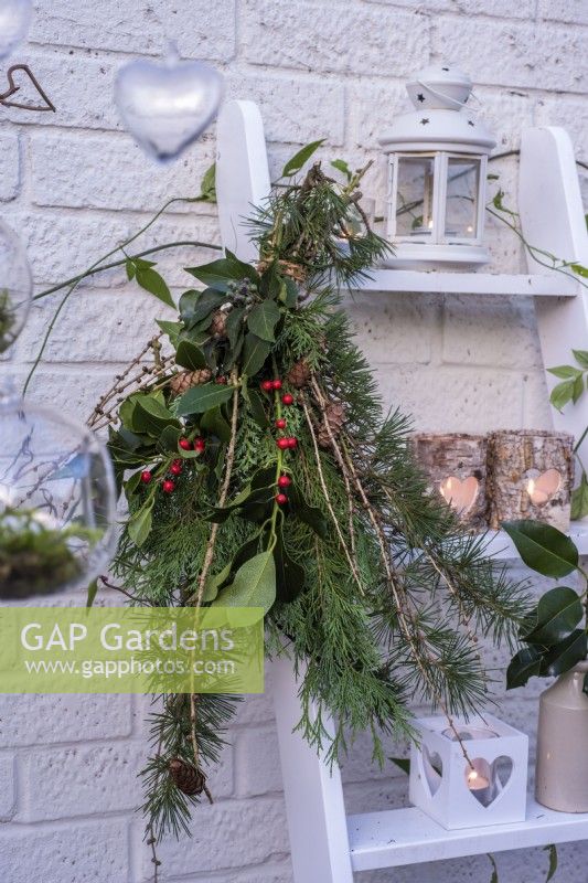 Tied bunch of evergreen foliage - conifers, ilex and Larix decidua cones attached to white ladder decorated with tealights