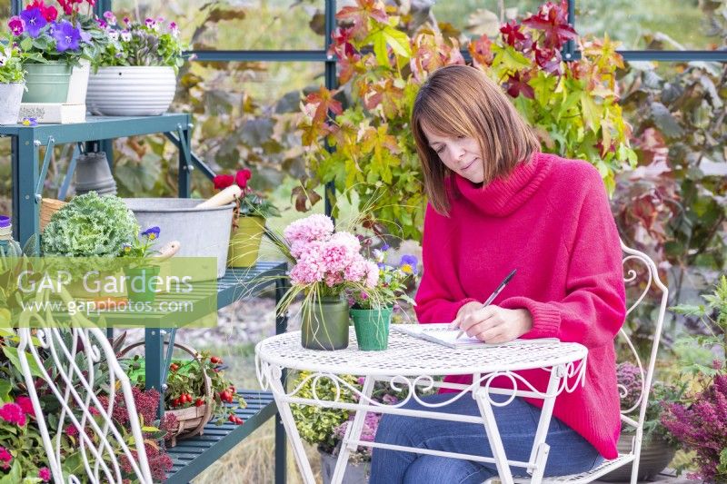 Woman sitting at a table writing in a greenhouse that has been filled with various plants and mixed containers