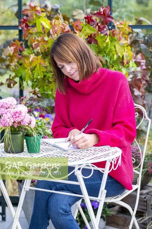 Woman sitting at a table writing in a greenhouse that has been filled with various plants and mixed containers