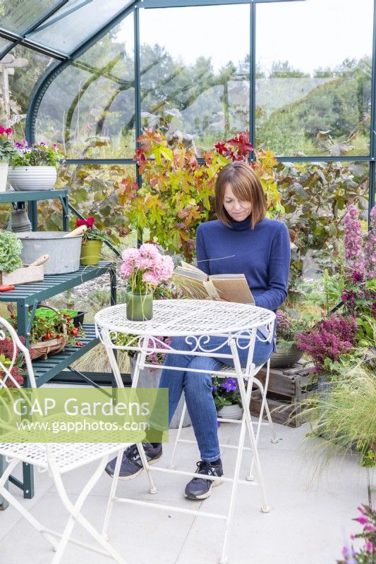 Woman sitting at a table reading in a greenhouse that has been filled with various plants and mixed containers