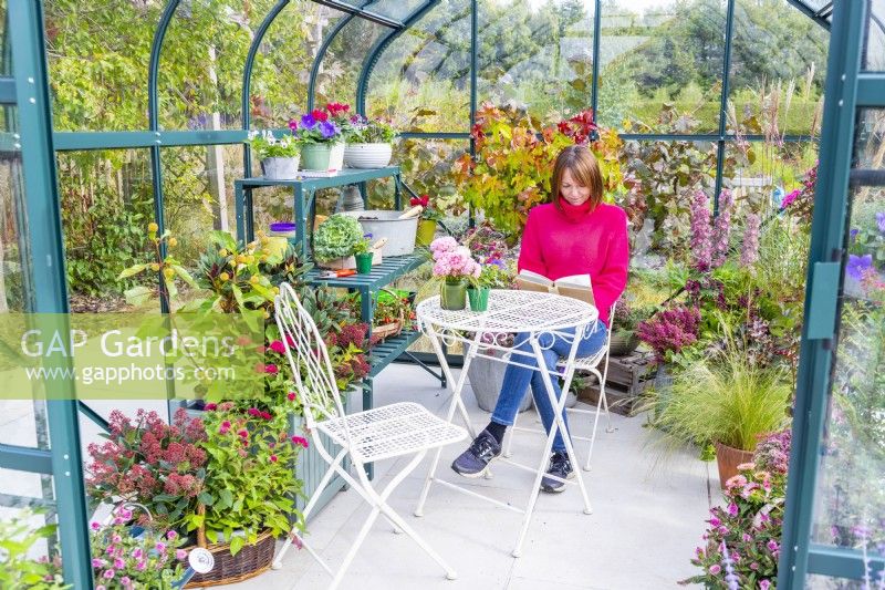 Woman sitting at a table reading in a greenhouse that has been filled with various plants and mixed containers