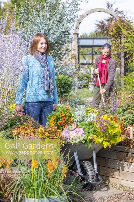 Woman pushing wheelbarrow full of plants through garden with woman sweeping in background