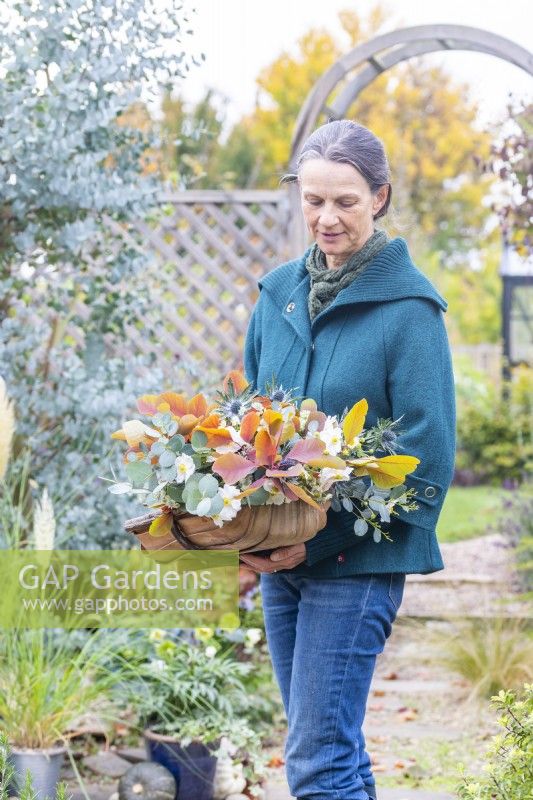 Woman holding an arrangement of Cotinus 'Grace', Eucalyptus 'Silver Dollar', Eryngium 'Picos Blue', Anemone 'Ruffled Swan' and Astrantia 'Snow Star' in a wooden trug