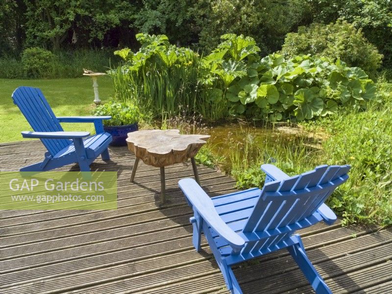 Painted wooden adirondack chairs and rustic table on decking overlooking natural garden pond