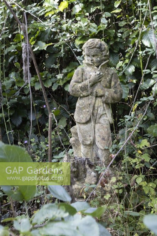 A weathered statue of a boy amongst overgrown and long grasses.  Autumn.