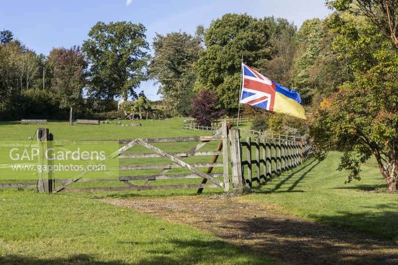 A gravel drive leads to a post and rail wooden gate and fence leading into a large grassy field. A Union Jack and Ukraine flag flies from the fence. Regency House, Devon NGS garden. Autumn