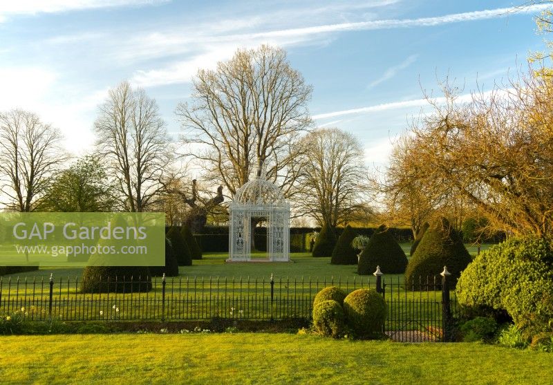 A white iron gazebo surrounded by pyramids of Taxus baccata on the Parterre Lawn at Chenies Manor.