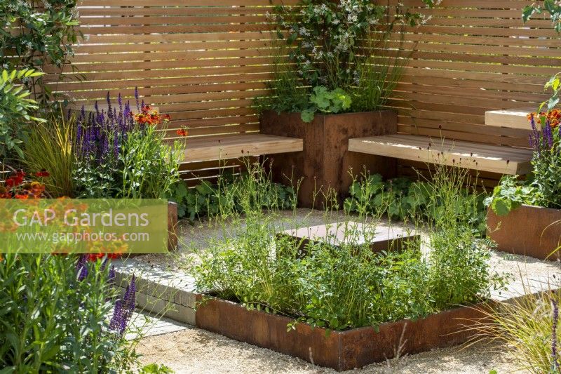 Seating area surrounded by wooden contemporary style fencing and Corten steel planters, hot planting includes Salvia 'Caradonna' and Helenium 'Moerheim Beauty' - The Lunch Break Garden, RHS Hampton Court Palace Garden Festival 2022