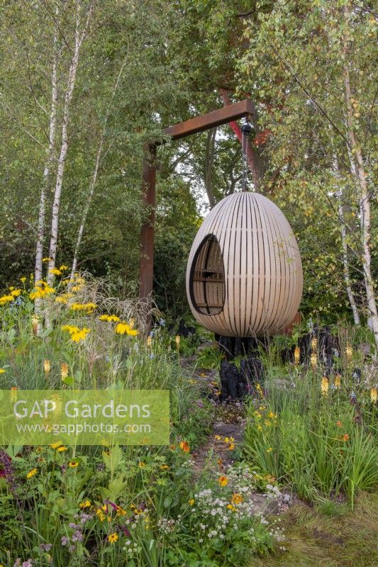 Bent oak swinging hide, suspended over naturalistic style planting, including Kniphofia, Rudbeckia 'Herbstonne' and Calamagrostis brachytrica - The Yeo Valley Organic Garden, RHS Chelsea Flower Show 2021