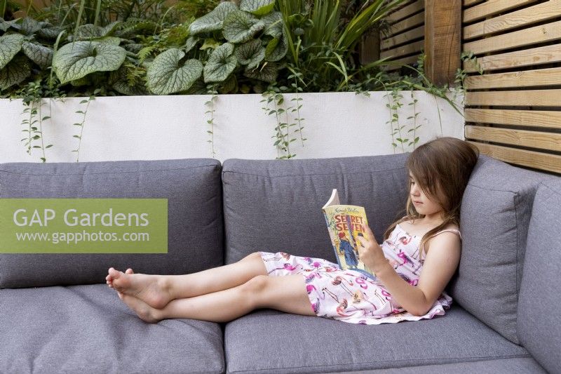 Young girl reading a book on garden seating with cushions.