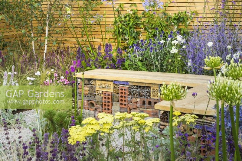 Bug or insect hotel inside gabion bench with wooden  top in low maintenance gravel garden with drought tolerant plants to attract pollinators, such as Salvia, Achillea, Agapanthus,Perovskia,  Stachys byzantina and Stipa tenuissima- Turfed Out, RHS Hampton Court Palace Garden Festival 202