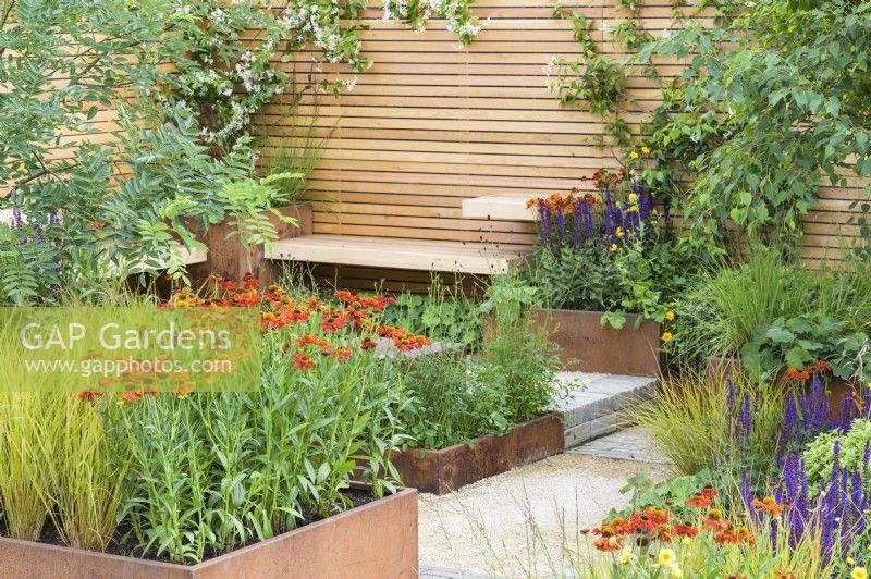 Rusted steel planters with perennials with  Helenium 'Moerheim Beauty' and simple wooden benches, fence and clay paver - Lunch Break Garden at RHS Hampton Court Palace Garden Festival 2022