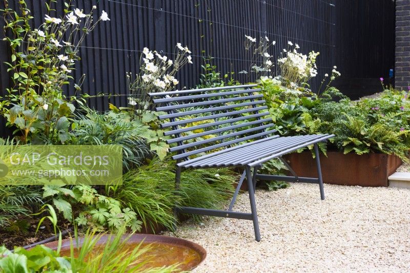 Black metal bench placed on gravel next to corten steel water bowl and the border with Anemone, Mahonia 'Soft Caress' and ferns by the black wooden fence. 