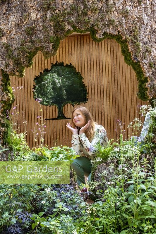 A woodland wildlife friendly garden with an oak tree trunk shelter and designer Taina Suuonio posing on the Connected by EXANTE Garden - RHS Chelsea Flower Show 2022 - Designed by Designer Taina Suonio - Built by Nicholsons 