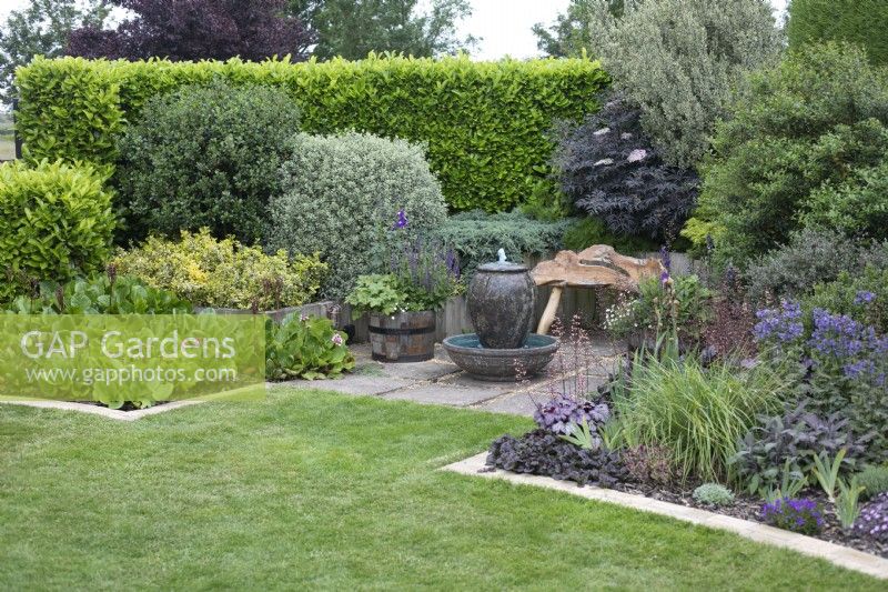 Seating area with water feature bordered by mixed beds of shrubs and summer flowers - June