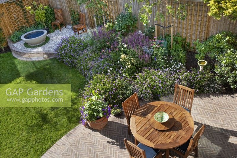 Overhead view of town back garden with circular herringbone brick paved patio, wooden table and chairs, small lawn, bowl water feature, herbaceous borders and new wooden boundary fencing