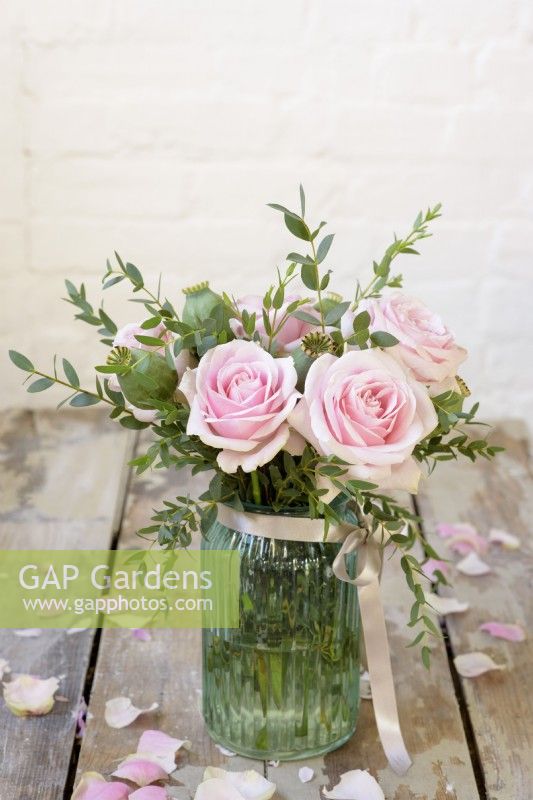 Pink roses arranged with eucalyptus and papaver seedheads in a blue glass jar tied with ribbon against rustic background