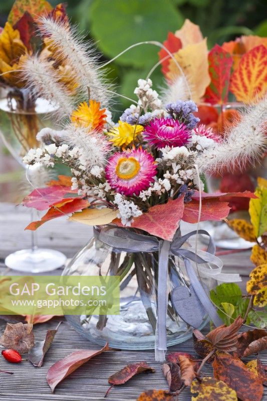 Flower bouqet containing strawflower, statice and grasses in glas vase decorated with autumnal leaves.