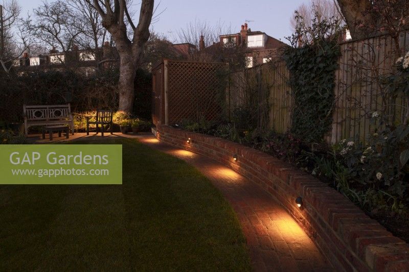 A London garden after a makeover with a new low brick wall with downlights along a brick path surrounded by lawn.