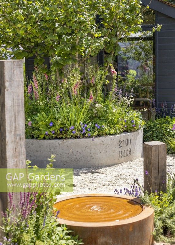 Contemporary water feature set in modern garden with upright wooden posts and conrete container with tree underplanted with perennials, summer June