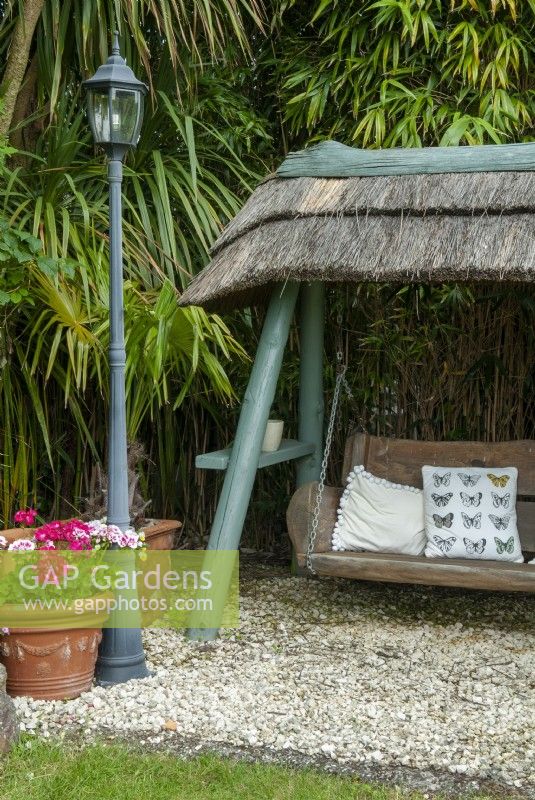 Garden recliner - swinging wooden seat under thatched roof against background of bamboo canes - Open Gardens Day, East Bergholt, Suffolk