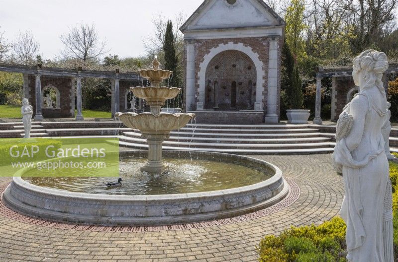 The entrance to the show gardens, with fountain, large, wisteria laden pergola and statues. A duck is swimming on the fountain pool. A classical stone statue is on the right. Brick paving surrounds the fountain pool. Trago Mills show gardens, Devon, UK. May. Spring