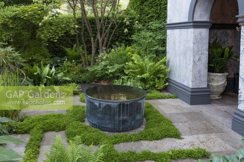 In a shady courtyard, a copper water container is encircled by baby's tears, Soleirolia soleirolii, planted in the cracks between paving slabs. 