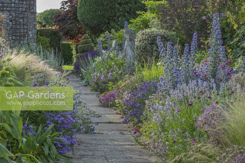 View of mixed perennials borders either side of a stone path in an informal country cottage garden in Summer - June