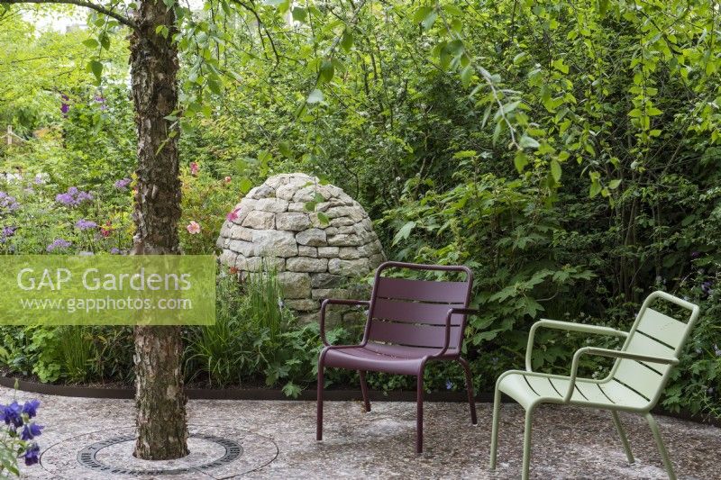 A tranquil shady seating area beneath Betula nigra, overlooked by a stone cairn immersed in a border with roses and thalictrum.