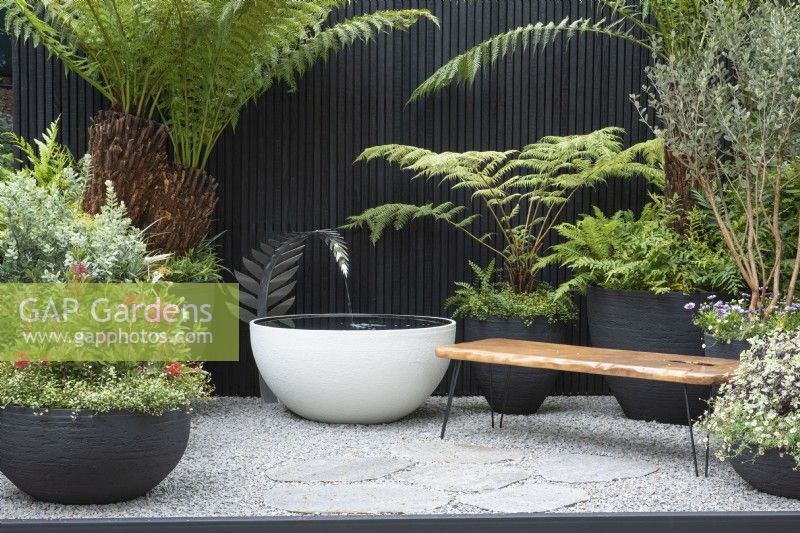 A courtyard inspired by New Zealand, with a timber seat made from reclaimed kauri wood, and handmade pots filled with native plants and tree ferns. A bespoke water feature is fed by a silver fern, NZ's national symbol.