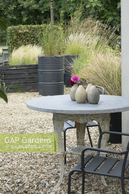 Gravel terrace. Classic iron table leg with marble top and old black painted barrels planted with several grasses. Dahlia in ceramic vase.