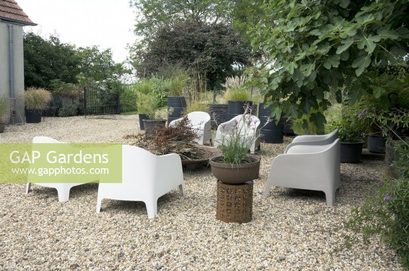 Terrace with gravel surface and seat area with metal shell fire pit. Container on decorated rustic iron table. Large fig tree and containers planted with several grasses.