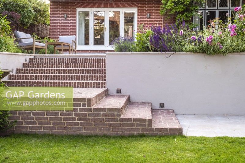 View of brick steps with recessed lighting and white rendered retaining wall leading up to raised terrace and house with mixed borders and furniture 