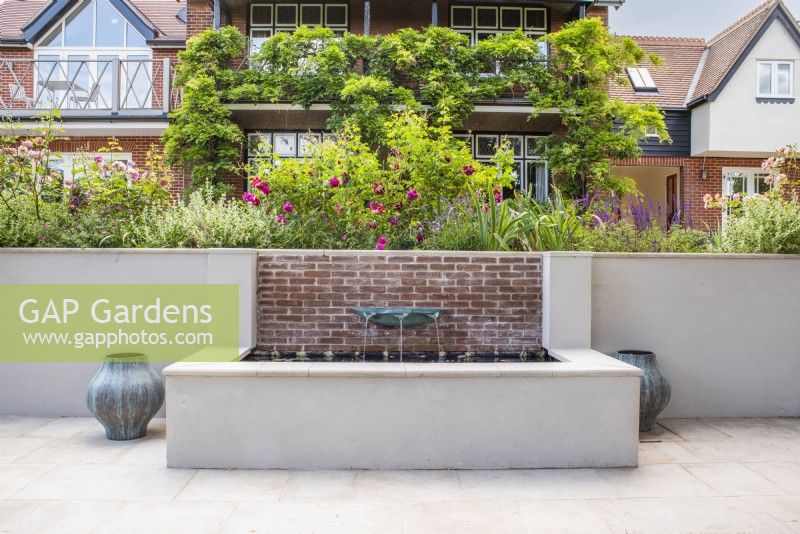 White rendered and brick raised water feature with verdigris basin fountain with containers either side and house in background