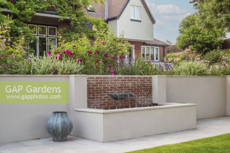 White rendered and brick raised water feature with verdigris basin fountain with verdigris containers and house and terrace borders in background