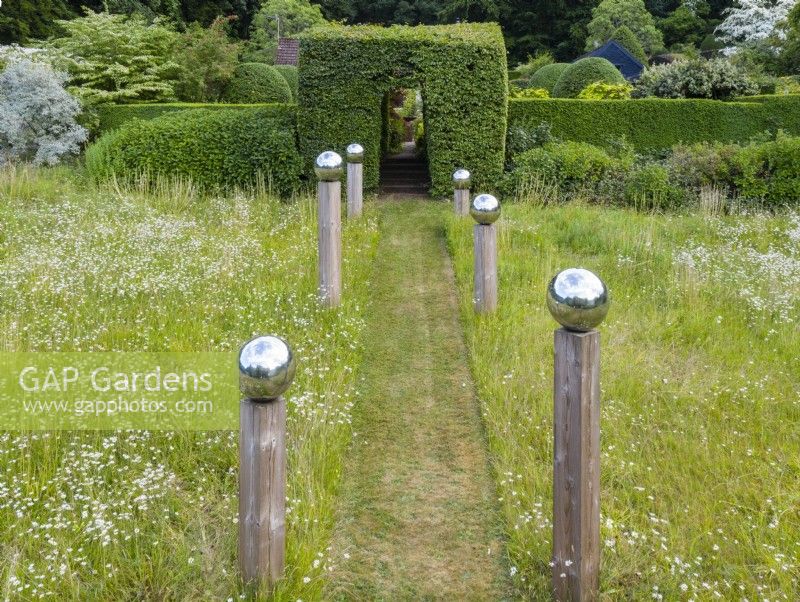 View over meadow with formal avenue of cut grass lined with wooden posts topped with stainless steel globes. Hedge of Box and large clipped hornbeam entrance June. Summer. Image taken with drone. 