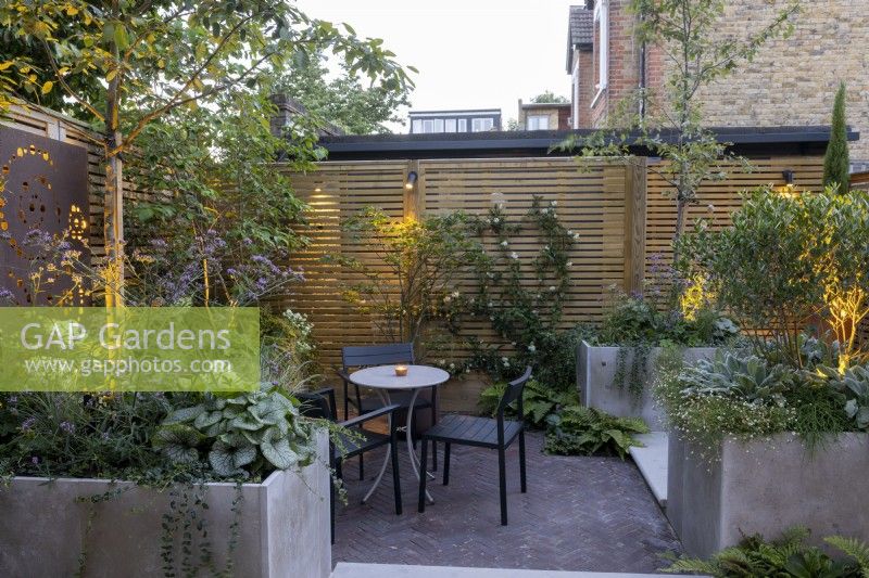 Courtyard garden at dusk with seating area, raised beds and contemporary wood boundary fence and lighting