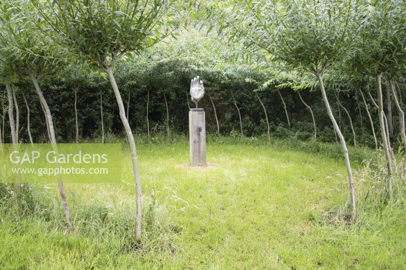 Palm of hand sculpture on wooden plinth in grass enclosure surrounded by willow standards. August. 