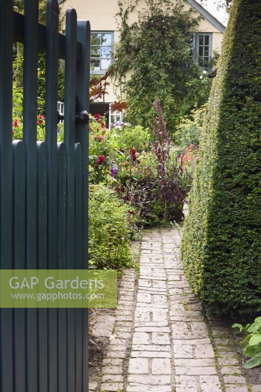 A gate opens onto a brick path leading into a country garden in August.
