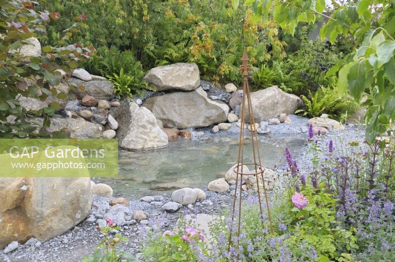An intimate small scale garden with large boulders and rocks surrounding a shallow central pool water feature with colour provided by perennials such as nepetas and roses.  The Oregon Garden - RHS Hampton Court Palace Garden Festival 2023.  Designed by Sadie May Stowell