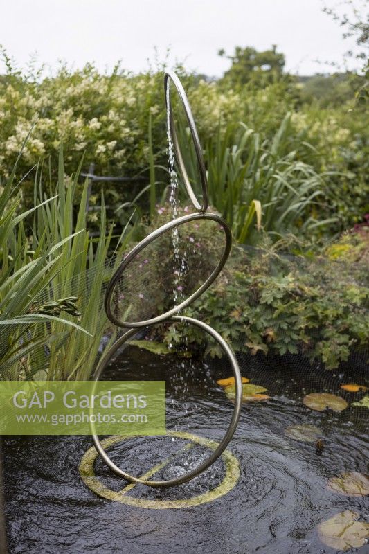 A rotating water feature, made of three interlinked hoops set in a pond. Water sprays downwards from the top hoop. A variety of water plants and other foliage is in the background. Harbour Lights, Devon NGS garden. July. 