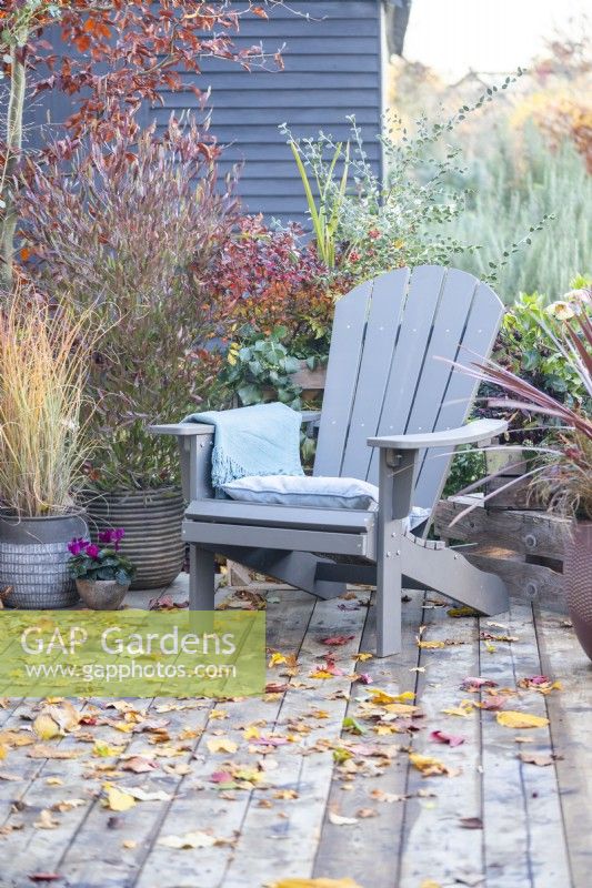 Recycled plastic chair with cushion and blanket surrounded by mixed planting and leaves scattered across a wooden deck